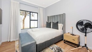 A modern bedroom with a large bed, bedside tables, a standing fan, and a view of the outdoors. - Spacious | Brand New | 2 Bed | Rosebank (Johannesburg)