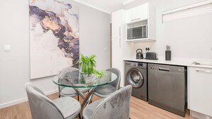 A modern kitchen with a glass dining table, grey chairs, a washing machine, a microwave, and a large abstract painting on the wall. - Spacious | Brand New | 2 Bed | Rosebank (Johannesburg)