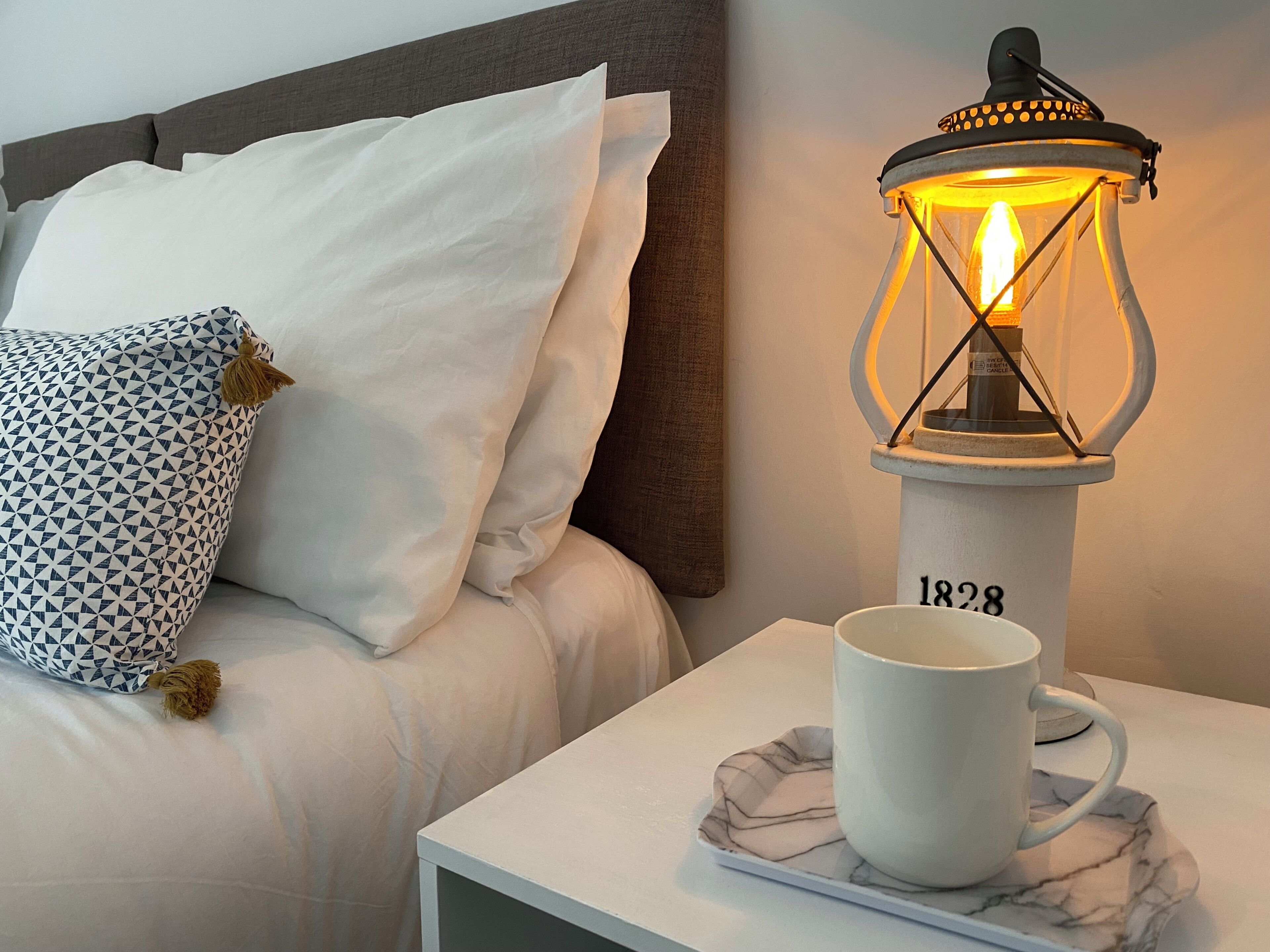 A bed with white bedding, a patterned pillow, a nightstand with a white mug, and a lit lantern.