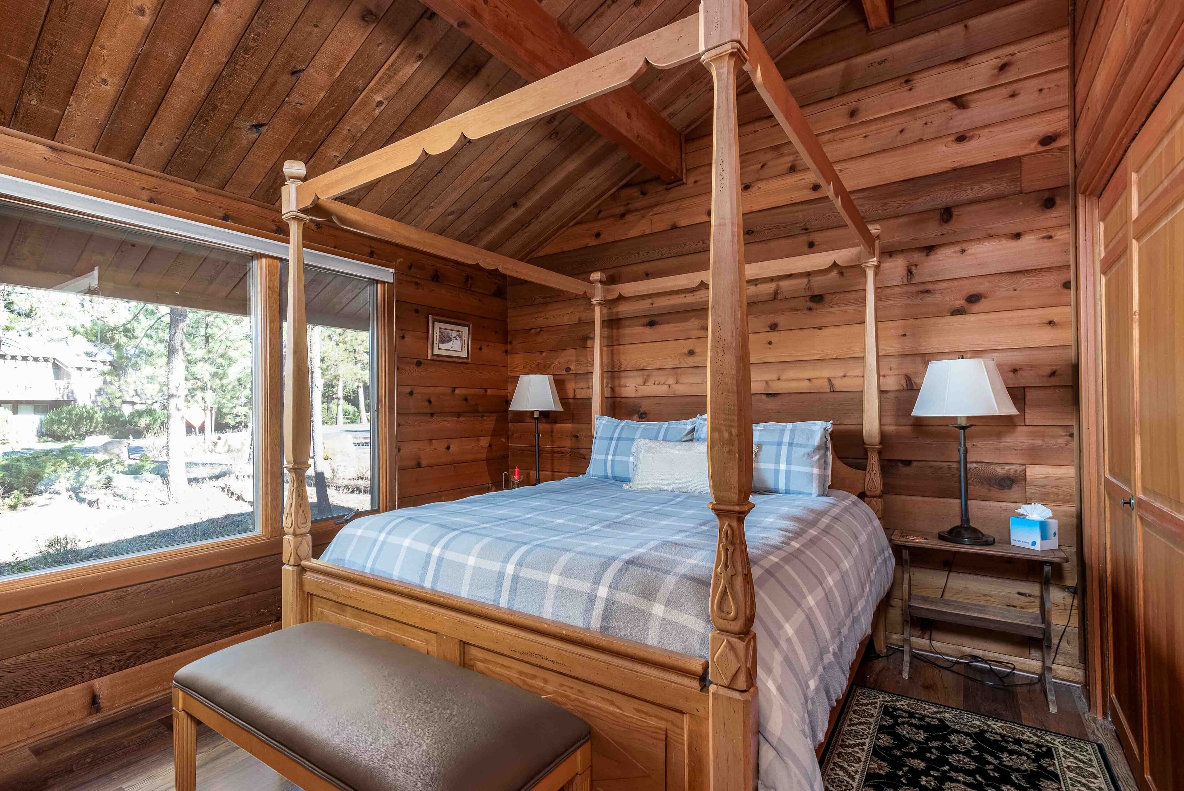 A wooden four-poster bed with a bench in front, a bedside table with a lamp, and a window with a view of the outdoors.
