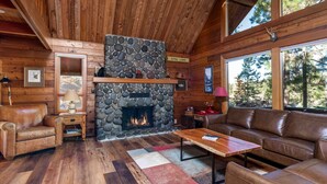 Interior acogedor de una cabaña con chimenea de piedra, muebles de cuero y grandes ventanales que dan a un bosque circundante.