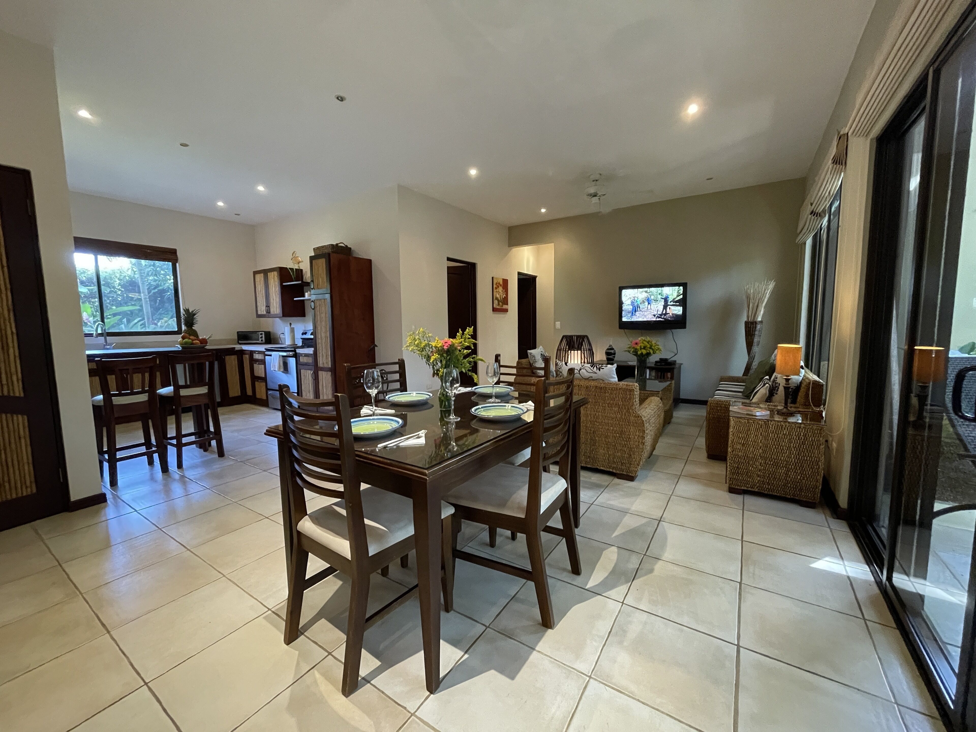 A spacious dining area with a large table and chairs, a kitchen in the background, and a television mounted on the wall.