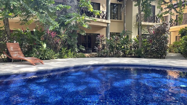 A swimming pool with clear blue water, surrounded by lush greenery and a wooden lounge chair. - 2 BR Condo Centrally Located in Playas del Coco (Playas del Coco)