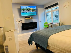 A hotel room with a bed, a TV mounted on the wall, a desk with a chair, and a window with a view of buildings.