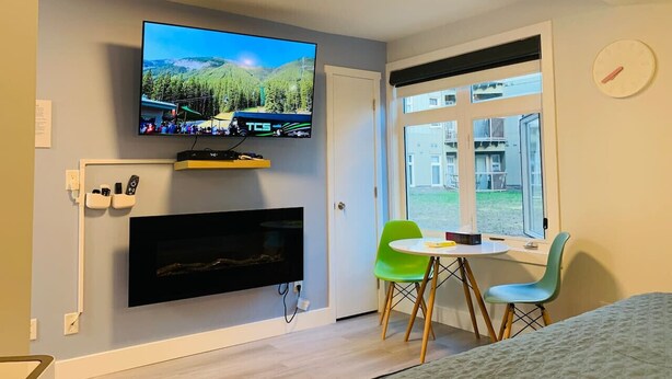 Una habitación con una televisión montada en la pared, una chimenea debajo, una mesa de comedor con sillas y un ventanal con vistas a edificios.