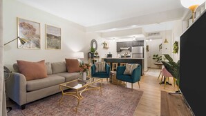 Ein modernes Wohnzimmer mit einer Couch, zwei Sesseln, einem Couchtisch und einem Fernseher. Im Hintergrund ist eine Küche mit Essbereich zu sehen.