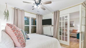 Ein Schlafzimmer mit einem großen Bett, einem Deckenventilator, einem Fernseher und einer Glastür, die in einen Wohnbereich führt.