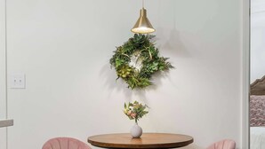 Ein Zimmer mit einem runden Holztisch, zwei rosa gepolsterten Stühlen mit goldfarbenen Beinen, einer hängenden Gold-Pendelleuchte und einem grünen Kranz an der Wand.