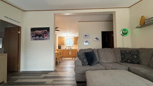 Ein Wohnzimmer mit einem grauen Ecksofa, Holzboden, einer Küche im Hintergrund und einer Wanddekoration mit Jeep-Motiv.
