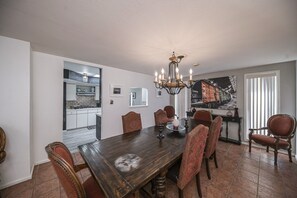 A dining room with a wooden table, six chairs, and a chandelier. There is a kitchen visible through an open doorway.