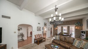 A spacious living room with a brown leather sofa, a wooden coffee table, and a chandelier. There is a fireplace with a brick wall behind it, and a dining area with a table and chairs. - Right At Home-Midland Adobe Compound with Pool and Fire Pits and Billiards (Midland)