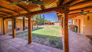 A covered patio area with wooden beams and tiled flooring, featuring a grassy lawn and a view of the surrounding landscape. - Right At Home-Midland Adobe Compound with Pool and Fire Pits and Billiards (Midland)