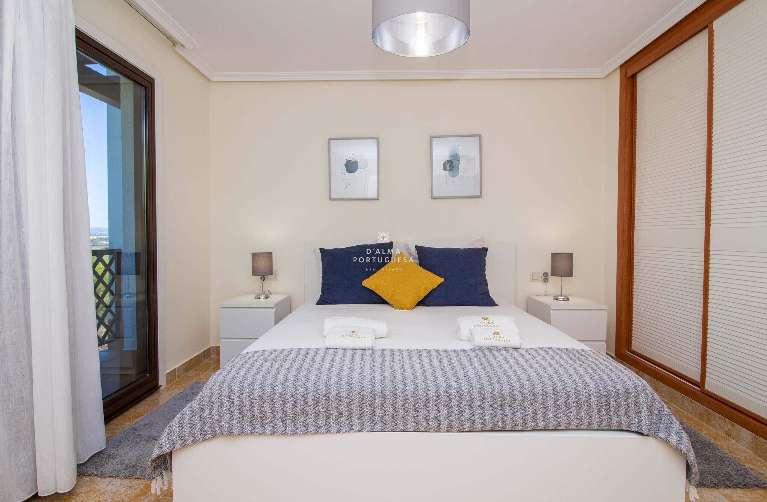 A hotel room with a bed, two bedside tables, a nightstand, a window with blinds, and a view of the outside.