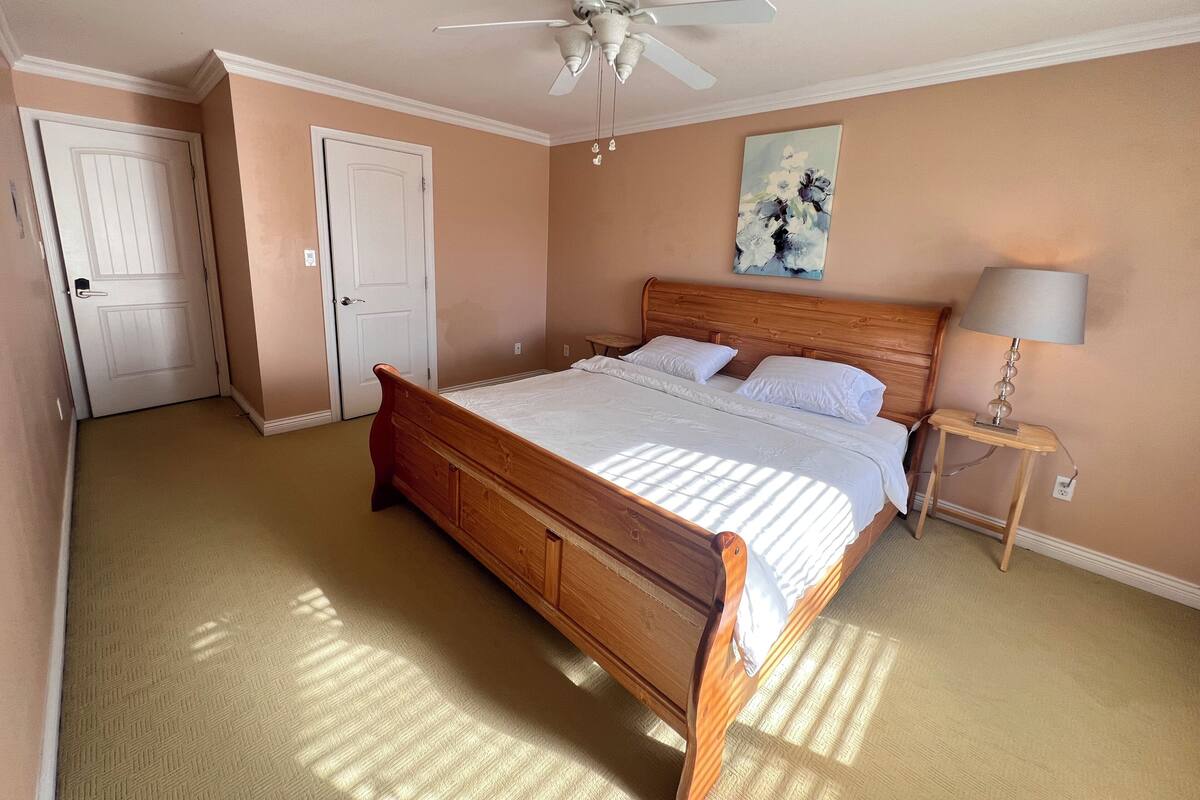 Un dormitorio con una cama de madera, un ventilador de techo, una mesita de noche con lámpara y un cuadro en la pared.