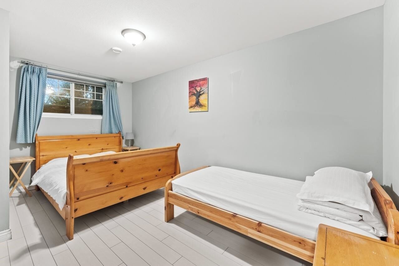 A room with two wooden beds, a window with curtains, a ceiling light, and a wall-mounted painting.