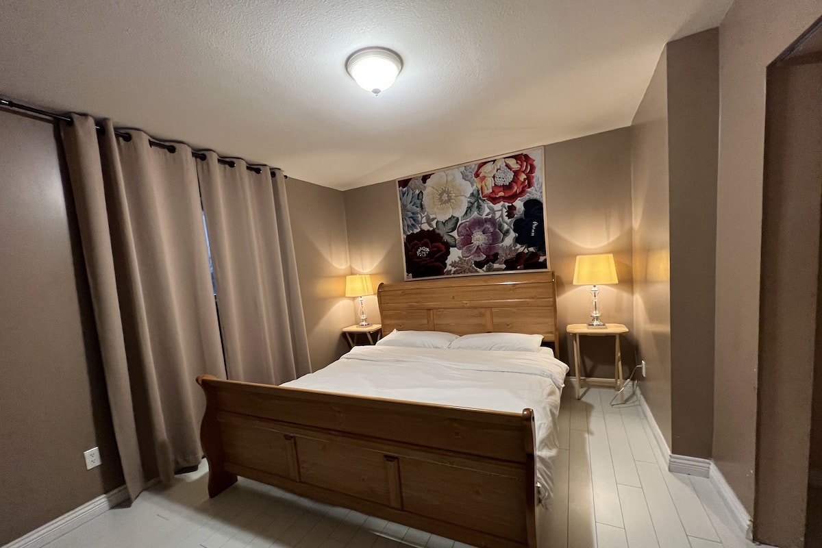 Habitación de hotel con una cama de madera, dos lámparas de noche, una obra de arte floral sobre la cama y cortinas color beige.