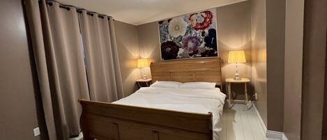 A hotel room with a wooden bed, two bedside lamps, a floral artwork above the bed, and beige curtains.