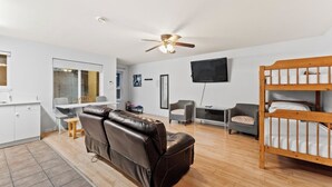 A modern, well-lit room with a ceiling fan, a wooden bunk bed, a leather sofa, a dining table with chairs, and a flat-screen TV mounted on the wall.