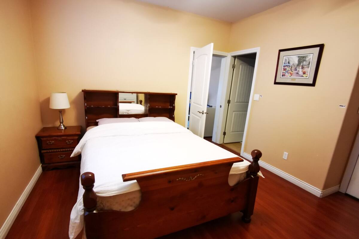 Un dormitorio con una cama de madera, una mesita de noche con una lámpara, una puerta que da a otra habitación y un cuadro en la pared.
