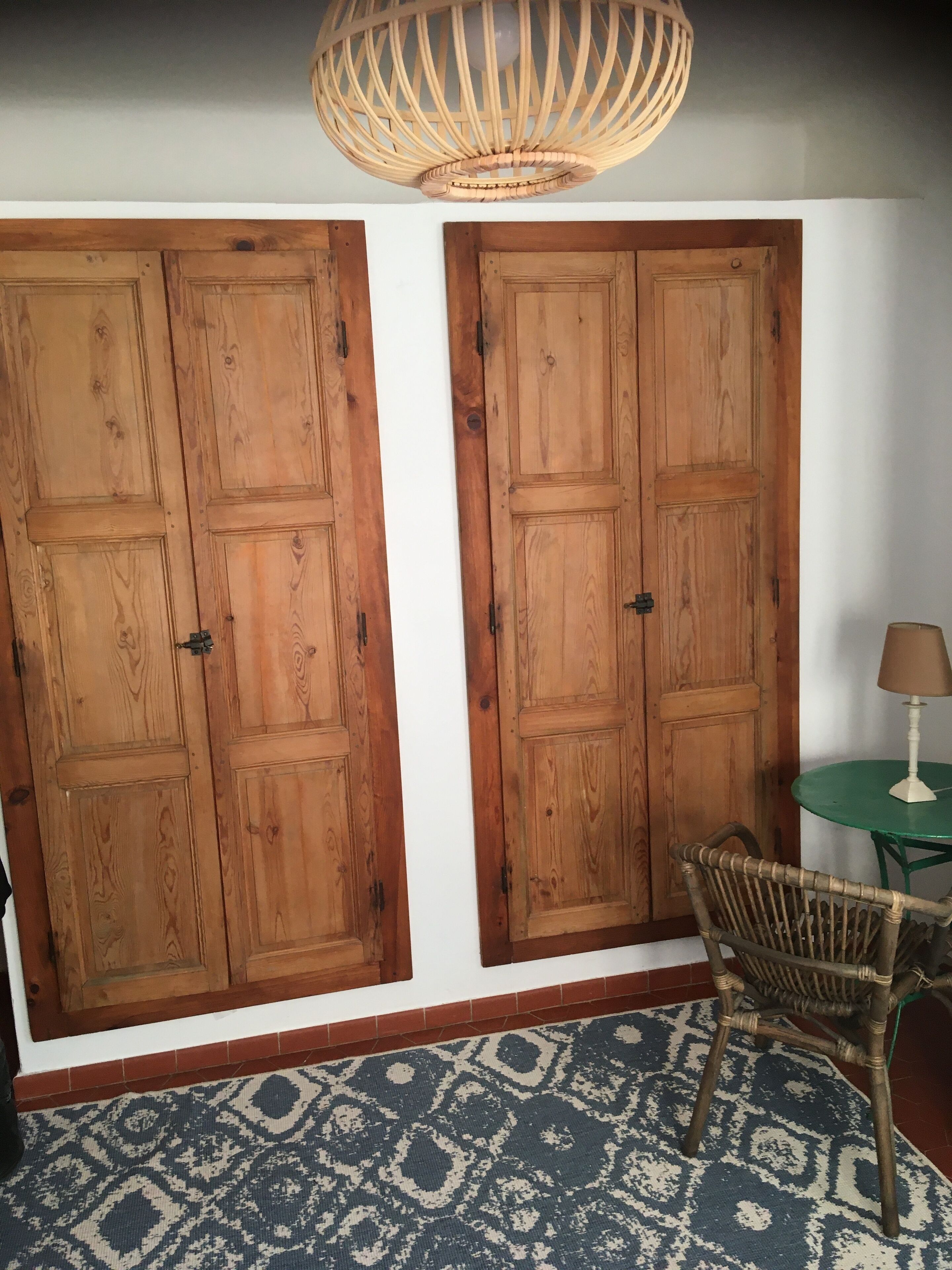 A room with two wooden wardrobes, a wicker chair, a small table with a lamp, and a patterned rug.