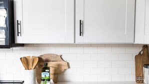 Una cocina con armarios blancos, una tabla de cortar de madera, una botella de aceite de oliva y un organizador de cucharas de madera.