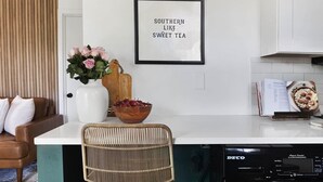 Una cocina con encimera blanca, un letrero enmarcado que dice 'SOUTHERN LIKE SWEET TEA', un jarrón con flores rosadas, una tabla de cortar de madera, un bol de cerezas, una silla de mimbre y un lavavajillas DECO.