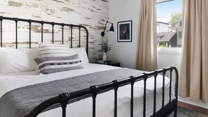 A bedroom with a bed, a black metal headboard, a bedside table with a lamp, a framed picture, and a window with curtains.