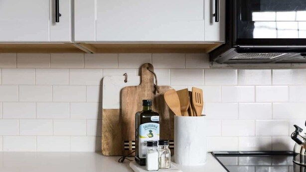 A kitchen with white cabinets, a wooden cutting board, a bottle of California Extra Virgin Olive Oil, salt and pepper shakers, and a white mug with utensils.