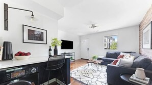 A modern living room with a sofa, a coffee table, and a TV. There is a kitchen area with a washing machine and a bar.
