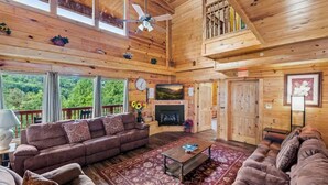 A spacious wooden cabin interior with a fireplace, large windows, and a ceiling fan. - HUGE 7BD Lodge IN TOWN +Pool Table + Views (Gatlinburg)