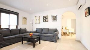 Un salon moderne avec un canapé gris, une table basse sur laquelle sont disposés des fruits, et un coin repas avec une table et des chaises blanches.