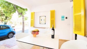 Une pièce avec une table blanche, des volets jaunes, une bouteille de vin et un saladier de fruits.