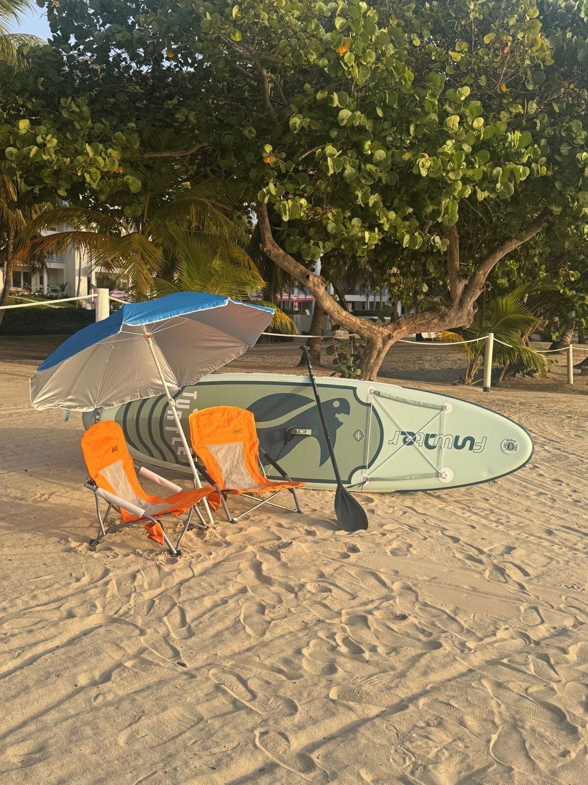 Escena de playa con una tabla de surf, dos sillas de playa y una sombrilla.