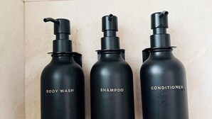Three black bottles with pump dispensers labeled 'BODY WASH,' 'SHAMPOO,' and 'CONDITIONER' against a tiled wall.