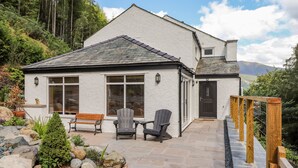 A white house with a stone roof, a wooden deck, and a stone pathway leading to the entrance. - Ladstock Cottage (Keswick)