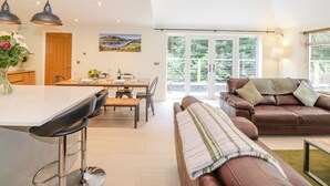 A modern living room with a brown leather sofa, a dining area with a wooden table, and a kitchen with pendant lights. - Ladstock Cottage (Keswick)