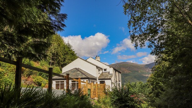 A house with a white exterior, surrounded by lush greenery and mountains in the background. - Ladstock Cottage (Keswick)