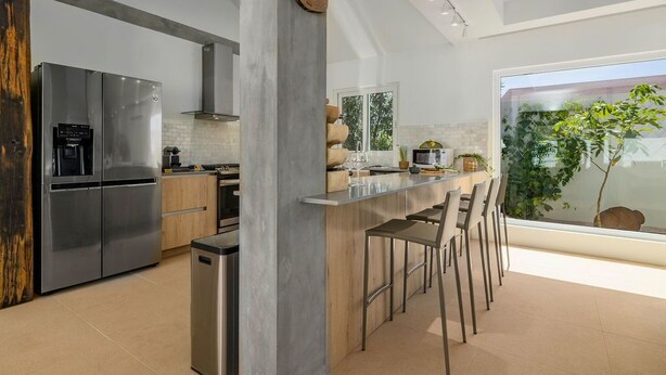 A modern kitchen with a stainless steel refrigerator, a wooden island with bar stools, and a large window offering a view of greenery. - Colonial Modern Villa Sleep 14-Pool, Palapa, Court (Noord)