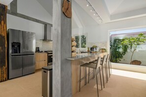 A modern kitchen with a stainless steel refrigerator, a wooden island with bar stools, and a large window offering a view of greenery. - Colonial Modern Villa Sleep 14-Pool, Palapa, Court (Noord)