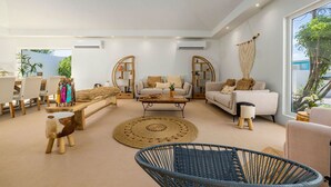 A spacious living room with a sofa set, a coffee table, and a wicker chair. There are two arch-shaped windows with curtains, and a large window on the right side. - Colonial Modern Villa Sleep 14-Pool, Palapa, Court (Noord)