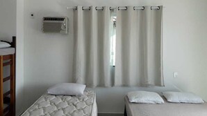 Una habitación con dos camas, una ventana con cortinas, un equipo de aire acondicionado y una silla de madera.