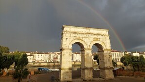 Ein uralter Steinbogen mit Inschriften, vor der Kulisse eines Flusses und Gebäuden, mit einem Regenbogen über der Szene.