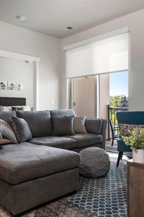 A modern living room with a grey sectional sofa, a round ottoman, and a patterned rug. - 2BR Corner Unit I Balcony + Trail Access + Bikes (Traverse City)