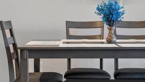 Une table à manger ornée d’un vase de fleurs bleues, deux chaises et un tableau représentant un coucher de soleil, accroché au mur.