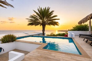 Ein Rooftop-Endlospool mit Blick auf das Meer bei Sonnenuntergang, umgeben von Liegestühlen und einer Palme.