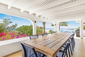 A covered outdoor dining area with a wooden table and chairs, surrounded by white pillars and a view of the sea. - Exclusive 10p Villa: Seaview, Pool & Private Beach (Bandabou)