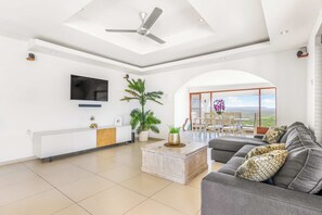 Ein geräumiges Wohnzimmer mit Deckenventilator, einem Flachbildfernseher, einer Couch, einem Couchtisch und Blick durch eine Glastür.