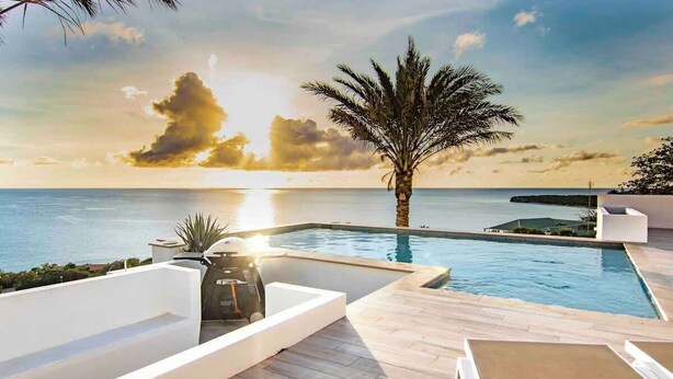 Un espace aménagé au bord de la piscine, offrant une vue sur la mer au coucher du soleil, avec un palmier et une architecture moderne.