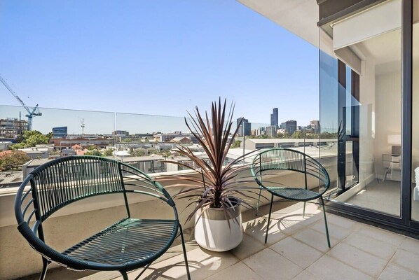 Enjoy the city skyline from this balcony with a glass door leading to the interior space, featuring two chairs and a potted plant.