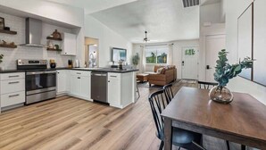 A modern kitchen with white cabinetry, a central island, and a dining area with a wooden table and chairs. - Nampa Greenbelt Retreat | Hot Tub | Walk to Indian Creek | Sleeps 10 (Nampa)
